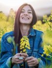 Délicieuse jeune brune en jean riant joyeusement démontrant des fleurs de colza jaune parfumées sur les mains debout sur le champ en fleurs le jour ensoleillé — Photo de stock