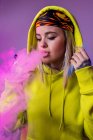 Впевнена жіноча хіпстерка в светрі курить електронну сигарету в студії на рожевому фоні і дивиться геть — стокове фото