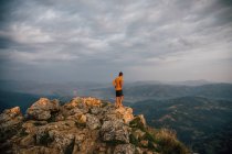 Homem distante em pé no topo da rocha acima do vale das terras altas sob o céu sombrio nas nuvens — Fotografia de Stock