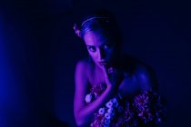 Attraktive junge barbusige Frau in weißen Hosen mit einem Strauß frischer bunter Blumen sitzt auf Hockern gegen grelles Neonlicht im dunklen Studio — Stockfoto
