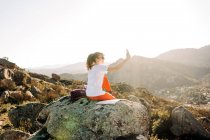 Vue arrière de jeune voyageuse heureuse aux longs cheveux foncés en vêtements décontractés et lunettes de soleil prenant selfie sur téléphone portable lors d'un voyage de randonnée en terrain montagneux — Photo de stock