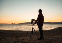 Вид збоку повне тіло невідомого чоловіка-фотографа, що стоїть біля штатива з фотокамерою і готується до зйомок морського пейзажу вдень — стокове фото