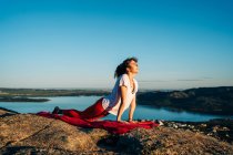 Side view of happy young female traveler in activewear smiling while practicing Upward Facing Dog with Neck Stretch yoga asana on rocky cliff under cloudless blue sky — Stock Photo