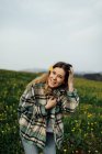 Content young female in checkered shirt looking at camera on meadow with blooming flowers under cloudy sky — Fotografia de Stock