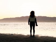 Back view silhouette of female traveler with photo camera standing contemplating seashore at sunset time — Foto stock