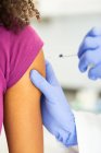 Especialista médica femenina irreconocible en uniforme protector y guantes de látex vacunando a una paciente afroamericana anónima en la clínica durante el brote de coronavirus - foto de stock