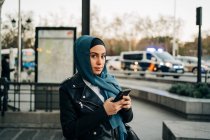 Encantadora musulmana con pañuelo en la cabeza de pie en la calle de la ciudad y navegar por el teléfono móvil mientras mira la cámara - foto de stock