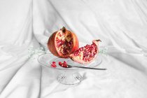 Яркий вкусный свежий гранат с спелыми семенами и цветущей веточкой цветов на прозрачной подставке на мятой ткани — стоковое фото