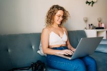 Молода щаслива самозайнята жінка в повсякденному одязі і окулярах сидить на зручному дивані і працює дистанційно на ноутбуці — стокове фото
