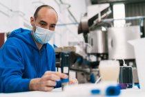 Male quality control specialist in sterile mask studying alcoholic liquid at table against professional equipment in factory — Stock Photo