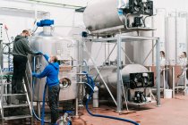 Seitenansicht männlicher Mitarbeiter mit Mundschutz, die während der Coronavirus-Pandemie Schlauch an Metallbehälter in Bierfabrik anschließen — Stockfoto