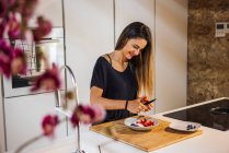 Zufriedenes Weibchen mit Messer, das reife Erdbeeren schneidet, während es zu Hause gesunde Nahrung in der Schüssel zubereitet — Stockfoto
