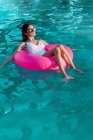 Молода весела жінка-мандрівник у купальнику та сонцезахисних окулярах лежить у надувному кільці в басейні — стокове фото