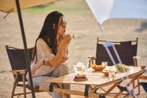Beautiful ethnic Asian female in sunglasses sitting at table drinking tea while having a relaxing time in camping area during holidays — Stock Photo