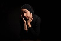Attraktive junge Islamistin in schwarzem Outfit und Hijab, die das Gesicht sanft berührt und nach unten schaut — Stockfoto