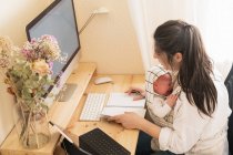 Adult mother sitting at desk working on desktop computer and taking notes in notebook while holding crying little child at table in daytime — Stock Photo