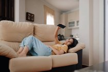 Seitenansicht einer jungen Frau beim Selbstporträt auf dem Handy, während sie sich im Wohnzimmer auf die Couch legt — Stockfoto