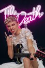 Energetische rebellische junge Frau in elegantem weißen Brautkleid und Kranz mit Gitarre in der Hand, die im Studio eine Horn-Geste mit Neonaufschrift macht — Stockfoto