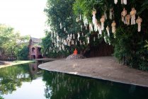Buda escultura en piedra bajo linternas en árboles contra la construcción oriental que refleja en estanque puro en Tailandia - foto de stock