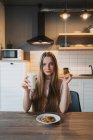 Веселая молодая женщина с вкусным овсяным печеньем с шоколадной крошкой на завтрак на столе на кухне — стоковое фото
