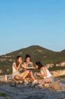 Companhia de jovens amigas viajando sentadas na colina e comendo ao pôr do sol em terras altas — Fotografia de Stock