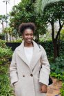 Fröhliche junge Afroamerikanerin in lässig warmem Mantel steht mit der Hand in der Tasche im grünen Stadtpark und blickt in die Kamera — Stockfoto