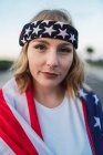 Retrato de encantadora mulher americana em bandana envolto em bandeira nacional dos EUA olhando para a câmera ao pôr do sol — Fotografia de Stock