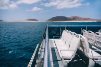Sedie bianche vuote sul ponte della barca da crociera che naviga nell'acqua blu del mare con la montagna sulla riva — Foto stock
