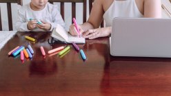 Cultiver femme méconnaissable et petit enfant assis à la table et dessin avec des marqueurs dans le carnet — Photo de stock