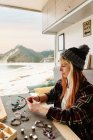 Вид збоку на вміст жінка мандрівник створює аксесуари ручної роботи, сидячи за дерев'яним столом у припаркованій вантажівці на морі — стокове фото