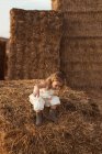 Curious little girl in overalls climbing down straw bale while playing in evening in countryside — Stock Photo