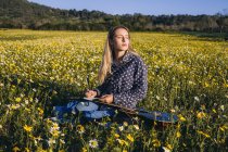 Молодая хипстерша, сидящая на лугу в сельской местности, пишет песни на блокноте и играет на гитаре во время летнего солнечного света. — стоковое фото