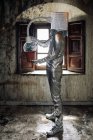 Side view unrecognizable person wearing silver costume with breathing apparatus and hose attached to potted plant standing in shabby abandoned room — Stock Photo