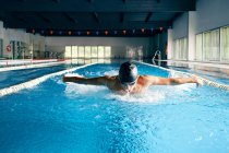 Strong male swimmer in bathing cap performing butterfly stroke during workout in swimming pool with blue water — Stock Photo