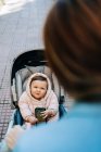 Desde arriba lindo bebé divertido con teléfono inteligente con ropa de abrigo sentado en el cochecito y mirando a la mamá borrosa en la calle de primavera - foto de stock