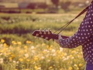 Vue arrière d'une jeune femme hipster méconnaissable qui se tient debout sur une prairie à la campagne en jouant de la guitare pendant la lumière du soleil d'été — Photo de stock