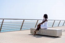 Vista lateral de longitud completa femenina afroamericana de moda en vestido blanco elegante sentado con las piernas cruzadas en el banco de piedra en el paseo marítimo de la ciudad y disfrutar de la vista al mar en el clima soleado - foto de stock