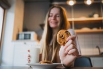 Alegre joven hembra con sabrosa galleta de avena con chips de chocolate para el desayuno en la mesa en la cocina - foto de stock