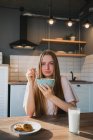 Junge Frau mit Löffel und Schale genießt leckere Maisringe, während sie in der Küche in die Kamera schaut — Stockfoto