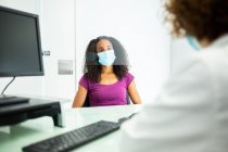 Afroamerikanerin mit Gesichtsmaske sitzt während eines Termins am zugeschnittenen, unkenntlich gemachten Schreibtisch einer Ärztin, die während des Coronavirus-Ausbruchs in einer modernen Klinik durch einen Plexiglasschirm geschützt ist — Stockfoto