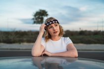 Young female in casual wear and headband with American flag print leaning out of car window while enjoying road trip at sunset — Stock Photo