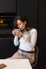 Содержание женщины с кружкой горячего напитка сидя на прилавке на кухне дома и глядя в сторону — стоковое фото
