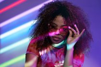 Portrait of attractive young African American female in stylish sunglasses looking at camera while standing in neon lights — Stock Photo