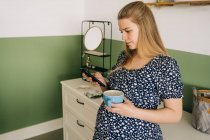 Joven mujer embarazada en ropa ornamental con taza de bebida caliente charlando en el teléfono celular en casa - foto de stock