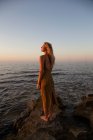 Vista lateral da jovem mulher em pé na costa contra o mar azul acenando ao pôr do sol e olhando para longe — Fotografia de Stock