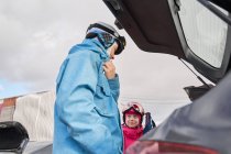 Vista lateral padre e hija usando ropa deportiva caliente y la colocación de esquís en el maletero del coche en el día de invierno soleado - foto de stock
