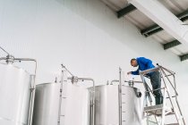 Engenheiro masculino na escada derramando líquido em tanque de aço inoxidável enquanto trabalhava na cervejaria — Fotografia de Stock