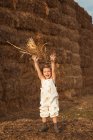 Fröhlich liebenswertes Kind in Overalls spielt mit Heu in der Nähe von Strohballen auf dem Land — Stockfoto