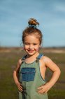 Encantada adorável menina em macacão de pé com as mãos na cintura no prado e olhando para a câmera — Fotografia de Stock