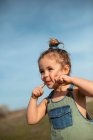 Délicieuse adorable petite fille en salopette debout avec les doigts sur le visage dans la prairie et regardant loin — Photo de stock